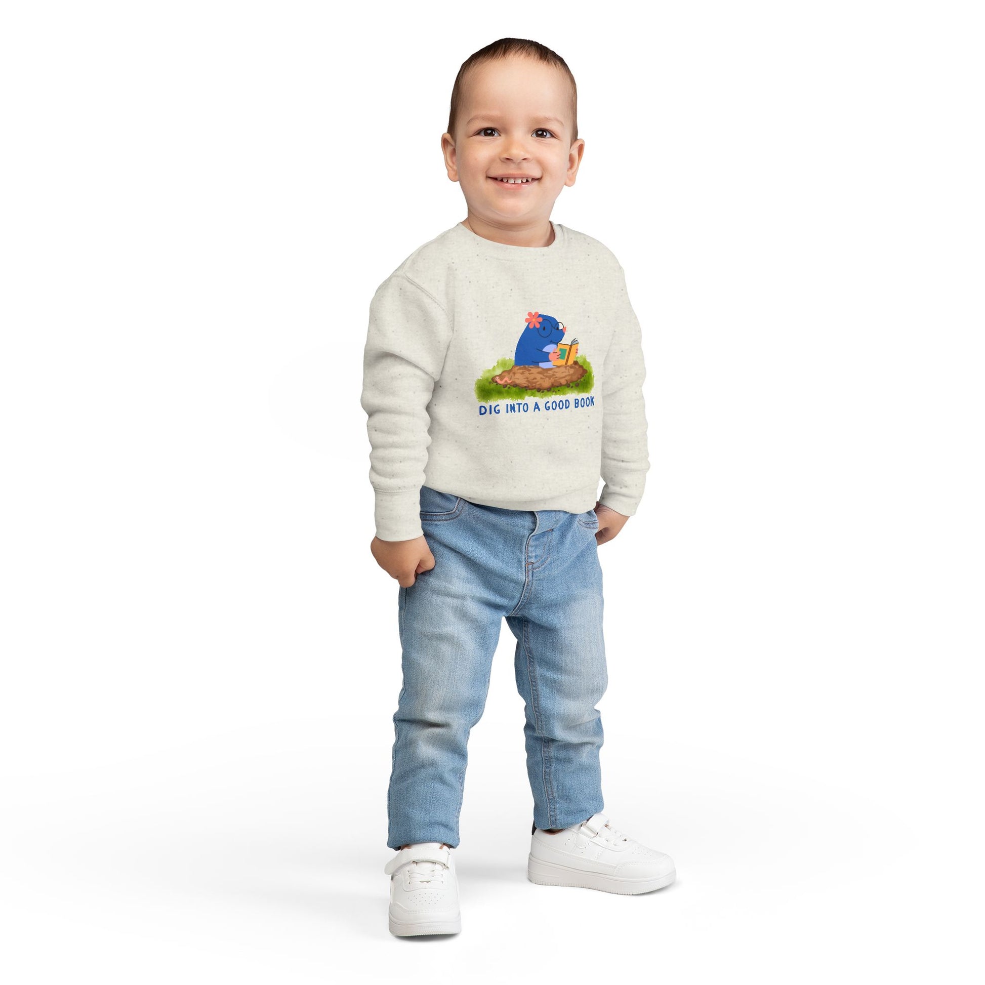 Boy wearing a beige toddler sweatshirt with a graphic of a mole reading a book and text 'Dig into a good book' 