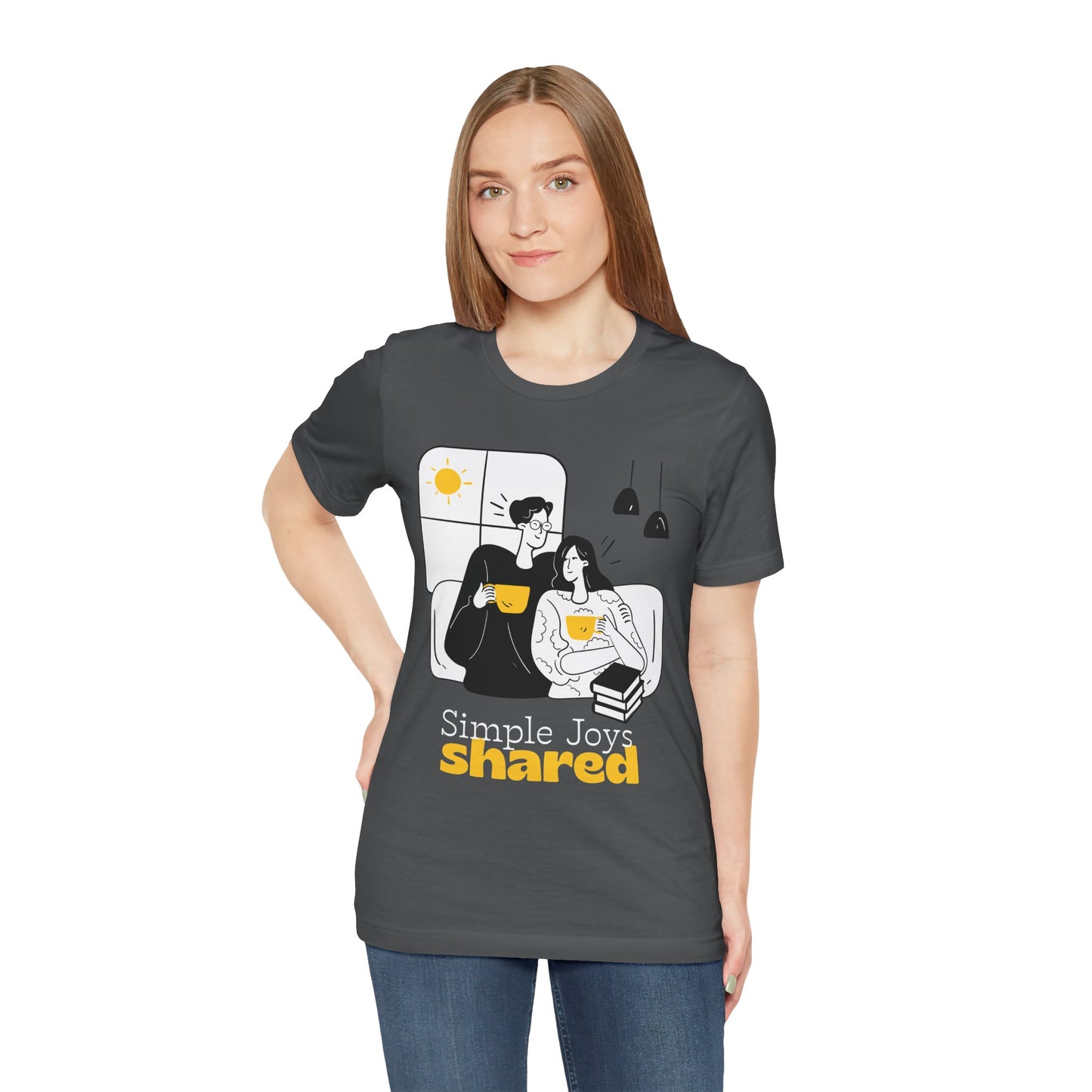 Woman wearing a gray t-shirt with a black, white and yellow graphic of a man an d woman snuggled together, drinking coffee, with a stack of books and the words 'Simple Joys Shared' text 