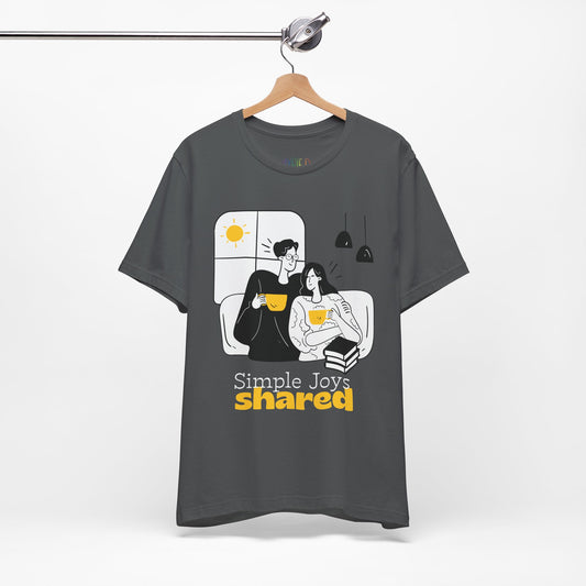 Gray t-shirt with a black, white and yellow graphic of a man an d woman snuggled together, drinking coffee, with a stack of books and the words 'Simple Joys Shared' text 