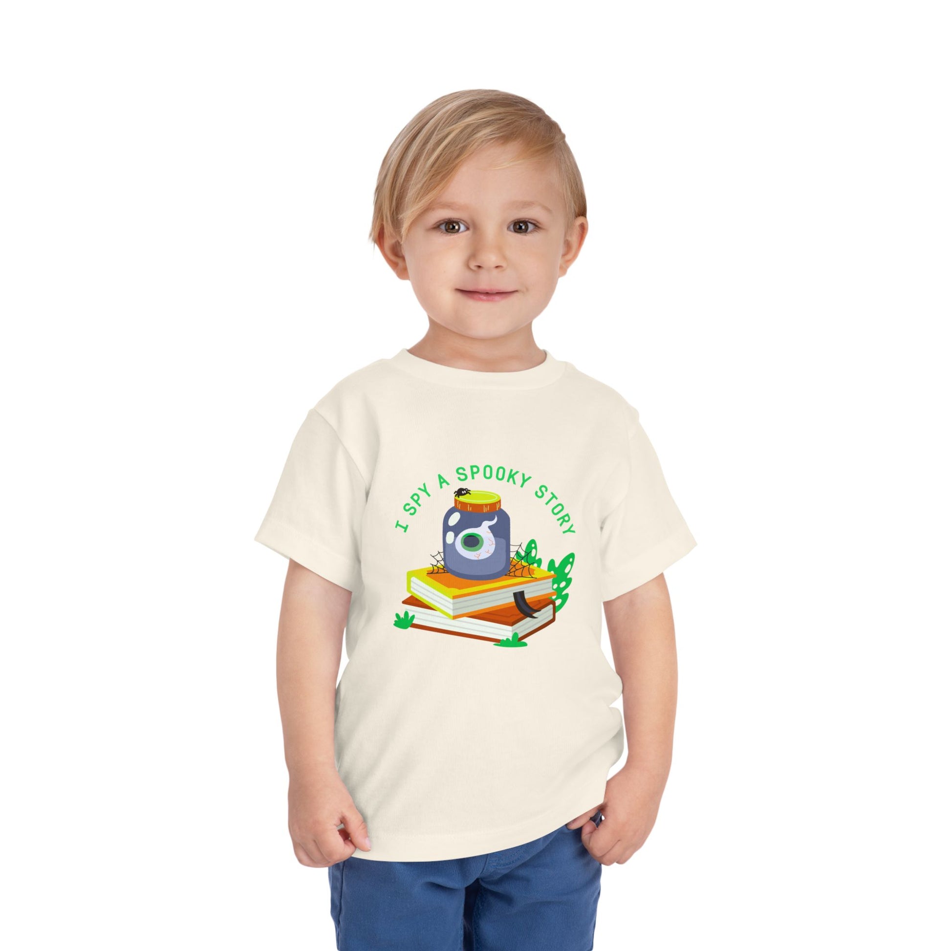 Child wearing a beige toddler t-shirt with an eyeball in a yar on top of books and text 'I spy a spooky story' on a white background
