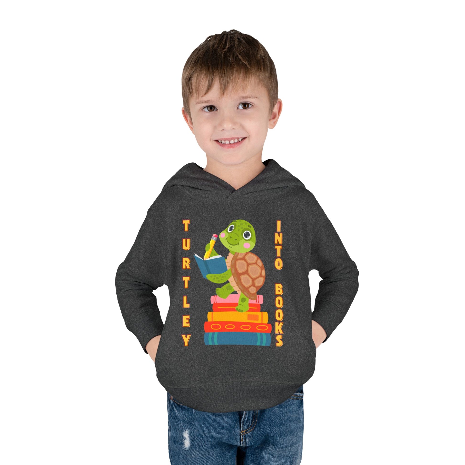 Boy wearing a dark gray toddler hoodie with a turtle graphic, notebook, stack of books and text 'Turtley Into Books'