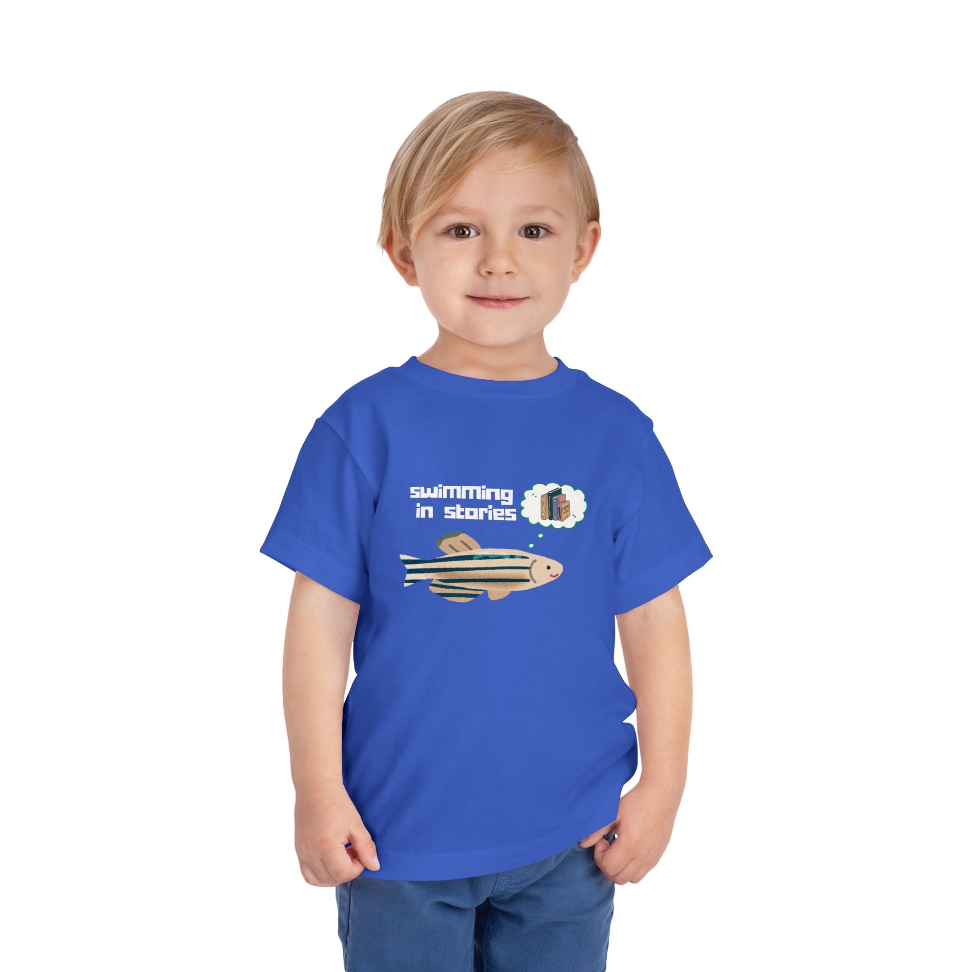 Toddler wearing a blue t-shirt with a fish graphic, thought bubble picturing books, and text 'swimming in stories' 