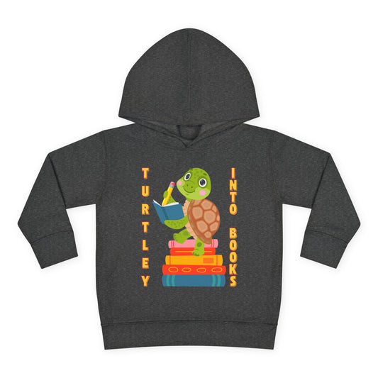 Smiling cartoon turtle perched on a stack of colorful books holding a pencil, with the words “Turtley Into Books” above — printed on a toddler hoodie.