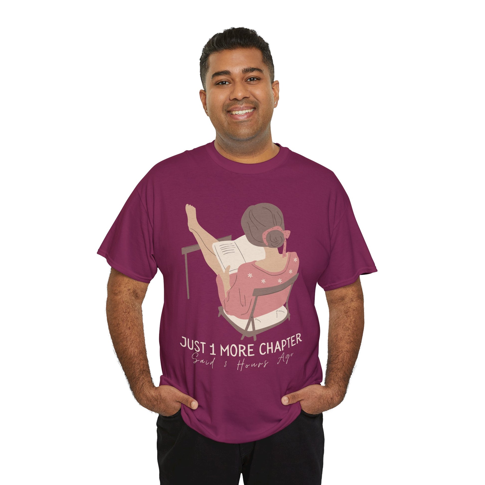 Man wearing a berry t-shirt with a graphic of a person reading a book with her feet propped up and text 'Just 1 More Chapter, Read's Hours Ago' 