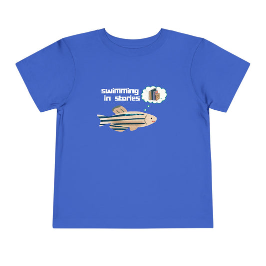 Blue t-shirt with a fish graphic, thought bubble picturing books, and text 'swimming in stories' 