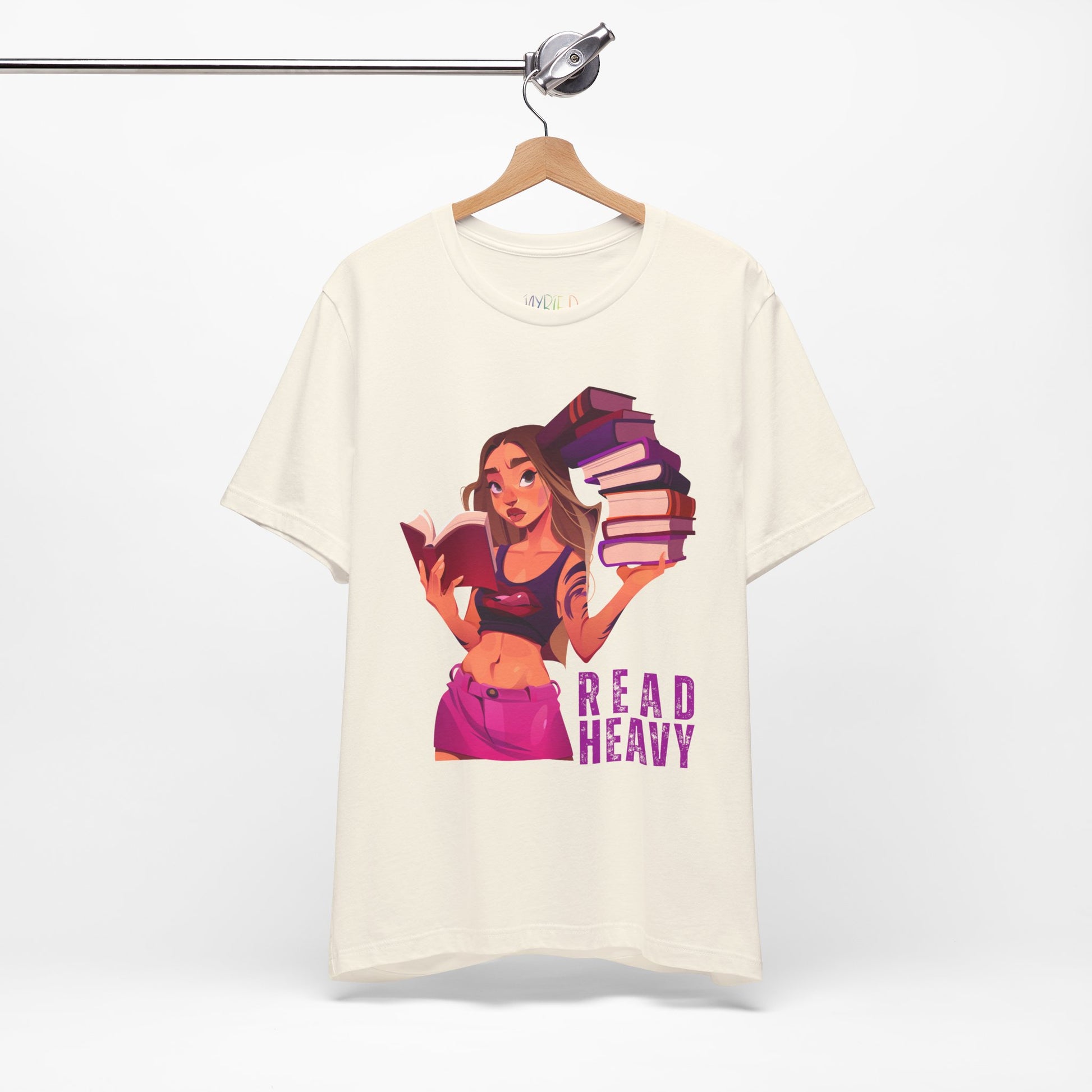 Beige t-shirt with a stylish woman graphic reading a book in one hand and holding a stack of books in the other, and the text 'Read Heavy' 