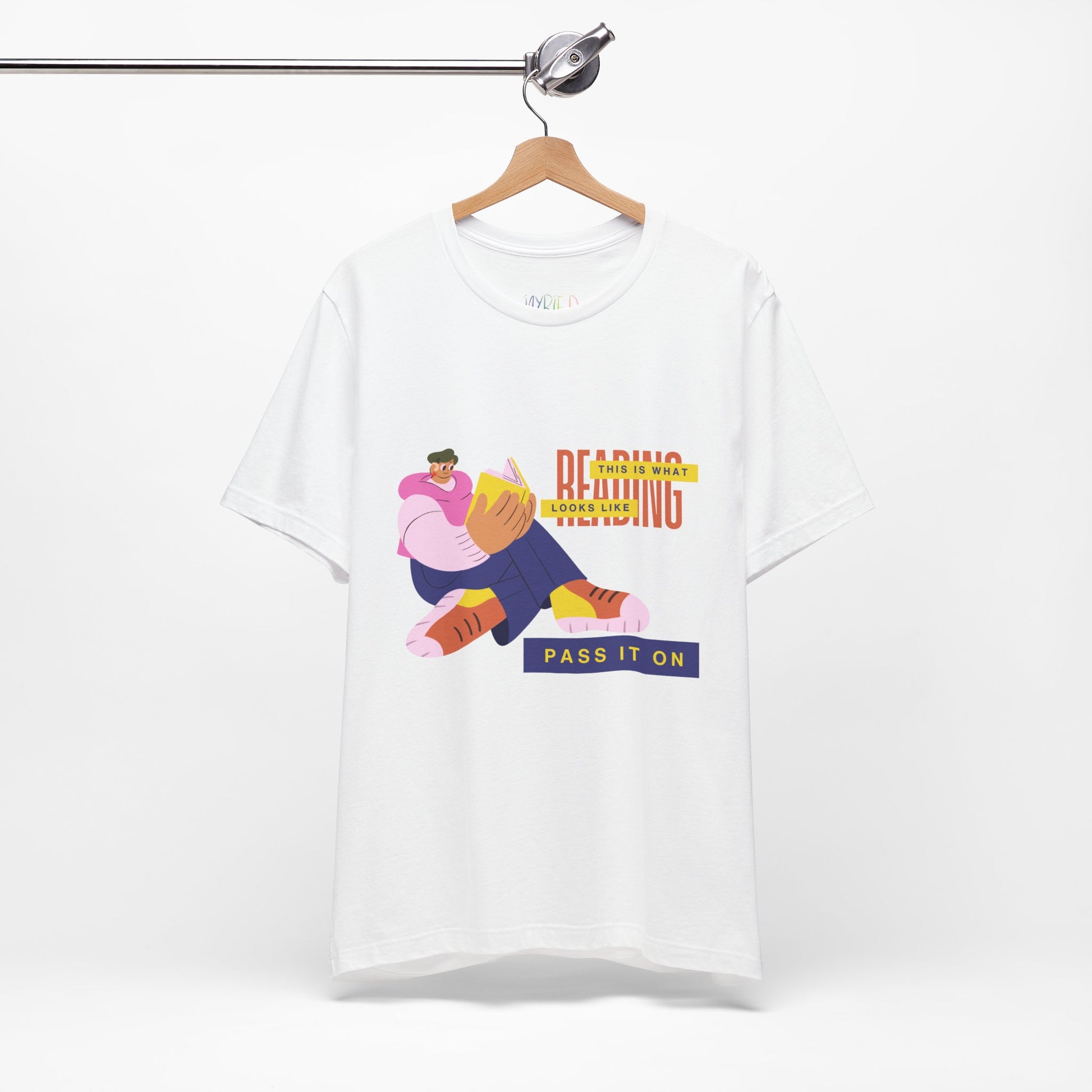 White t-shirt with colorful, funny shaped figure reading a book and text 'This Is What Reading Looks Like, Pass It On'