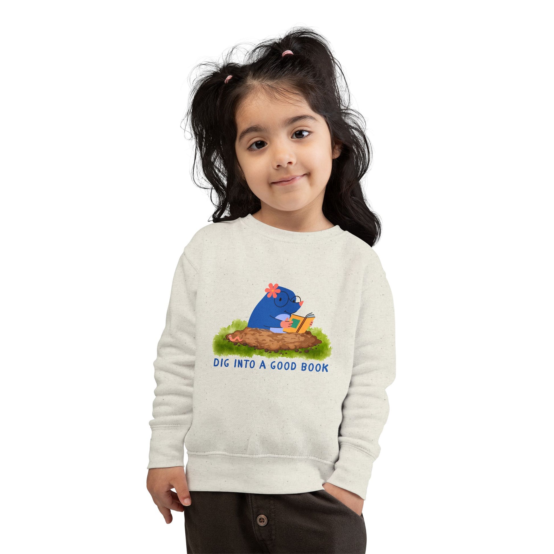 Girl wearing a beige toddler sweatshirt with a graphic of a mole reading a book and text 'Dig into a good book' 