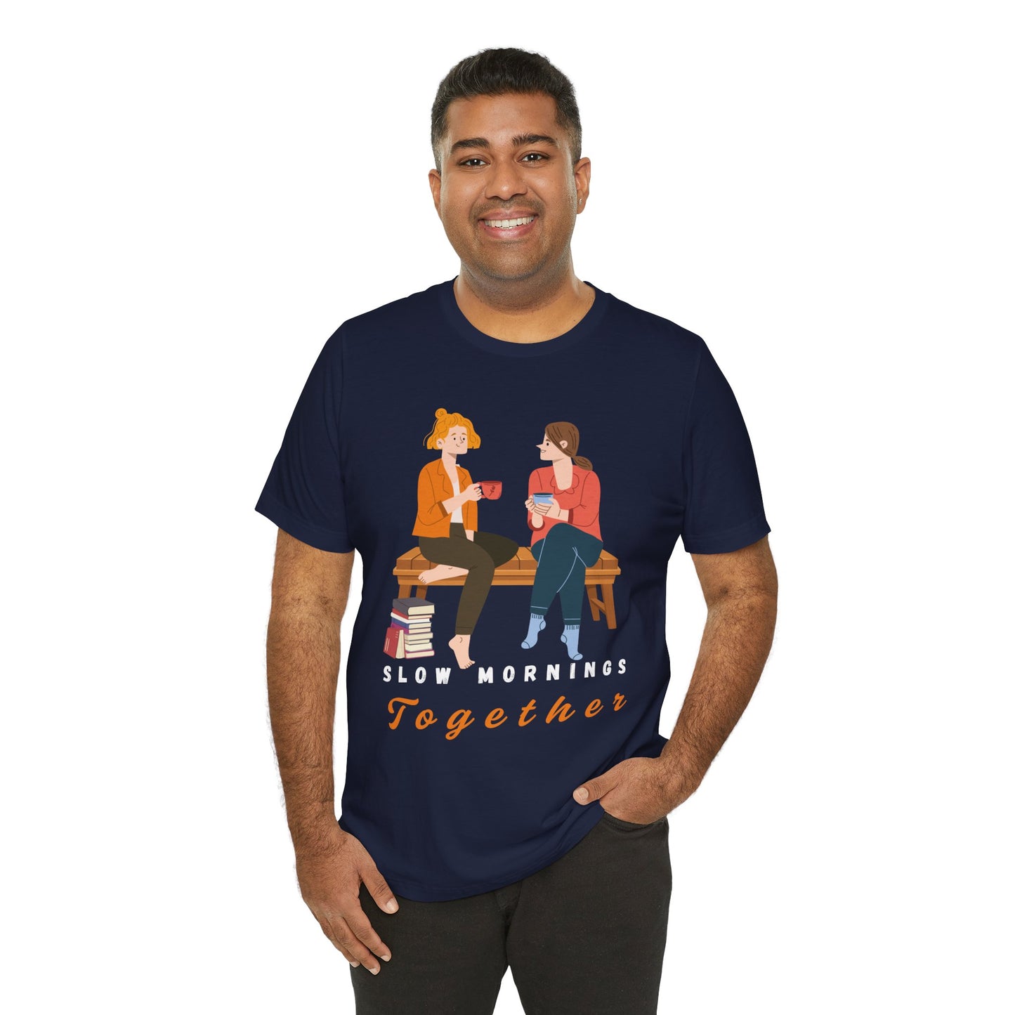 Man wearing a navy blue t-shirt with graphic of two people sitting on a bench together, hot beverages in their hands, book at their feet and 'Slow Mornings Together' text below the image