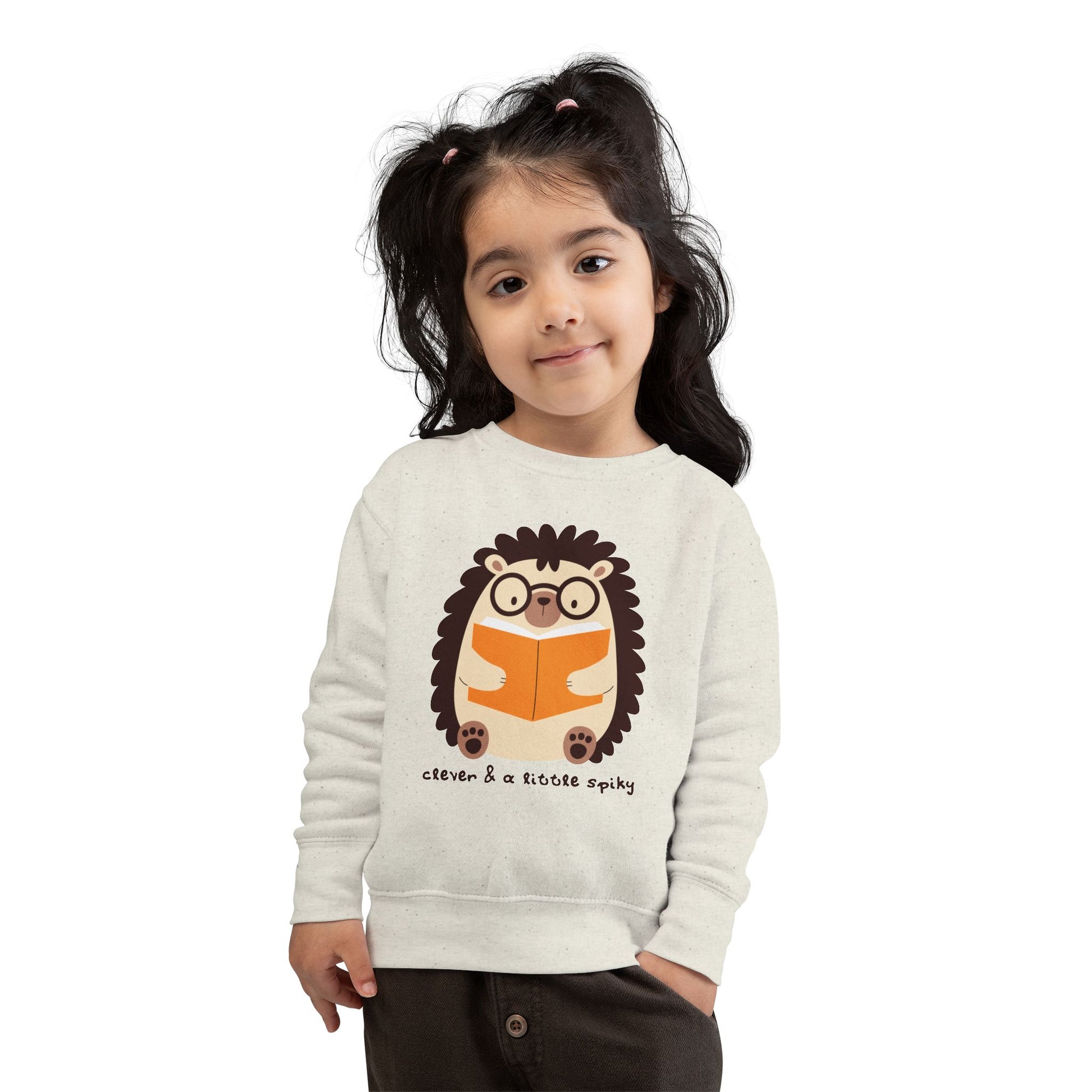 Girl wearing a beige toddler sweatshirt with a hedgehog graphic reading a book and text 'clever & a little spiky'