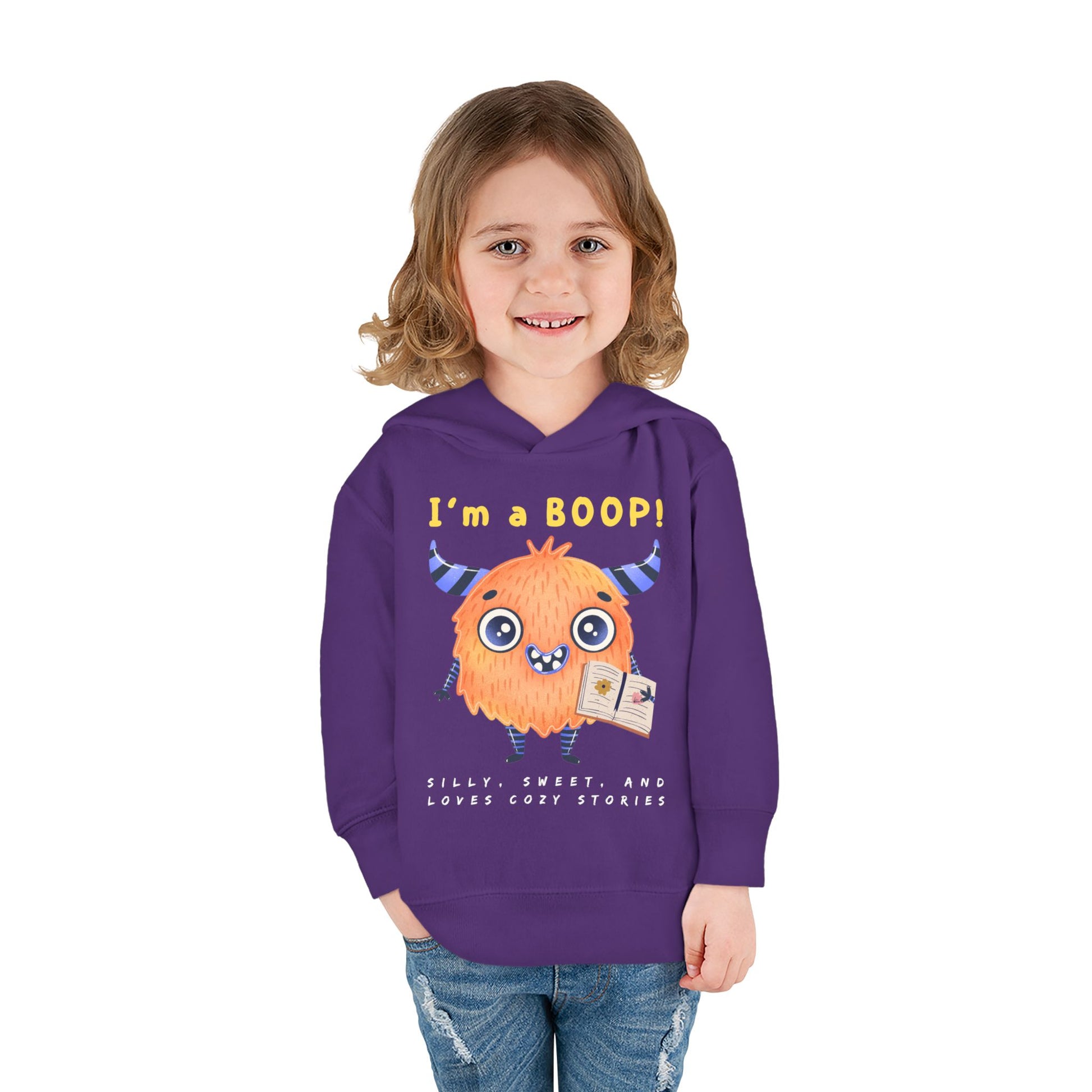 Girl wearing a purple toddler hoodie with a cartoon monster holding a book and text 'I'm a Boop', sub-text 'Silly, Sweet, And Loves Cozy Stories' on white background