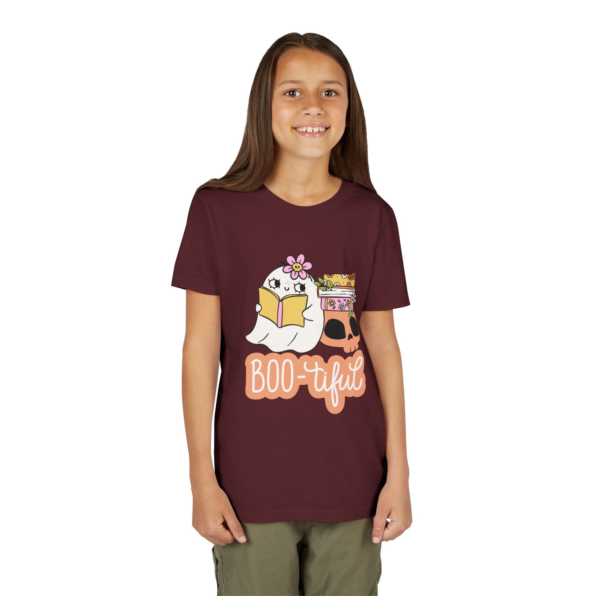 Girl wearing a maroon t-shirt with cartoon ghost leaning on a skull and stack of books while reading and text 'BOO-tiful'