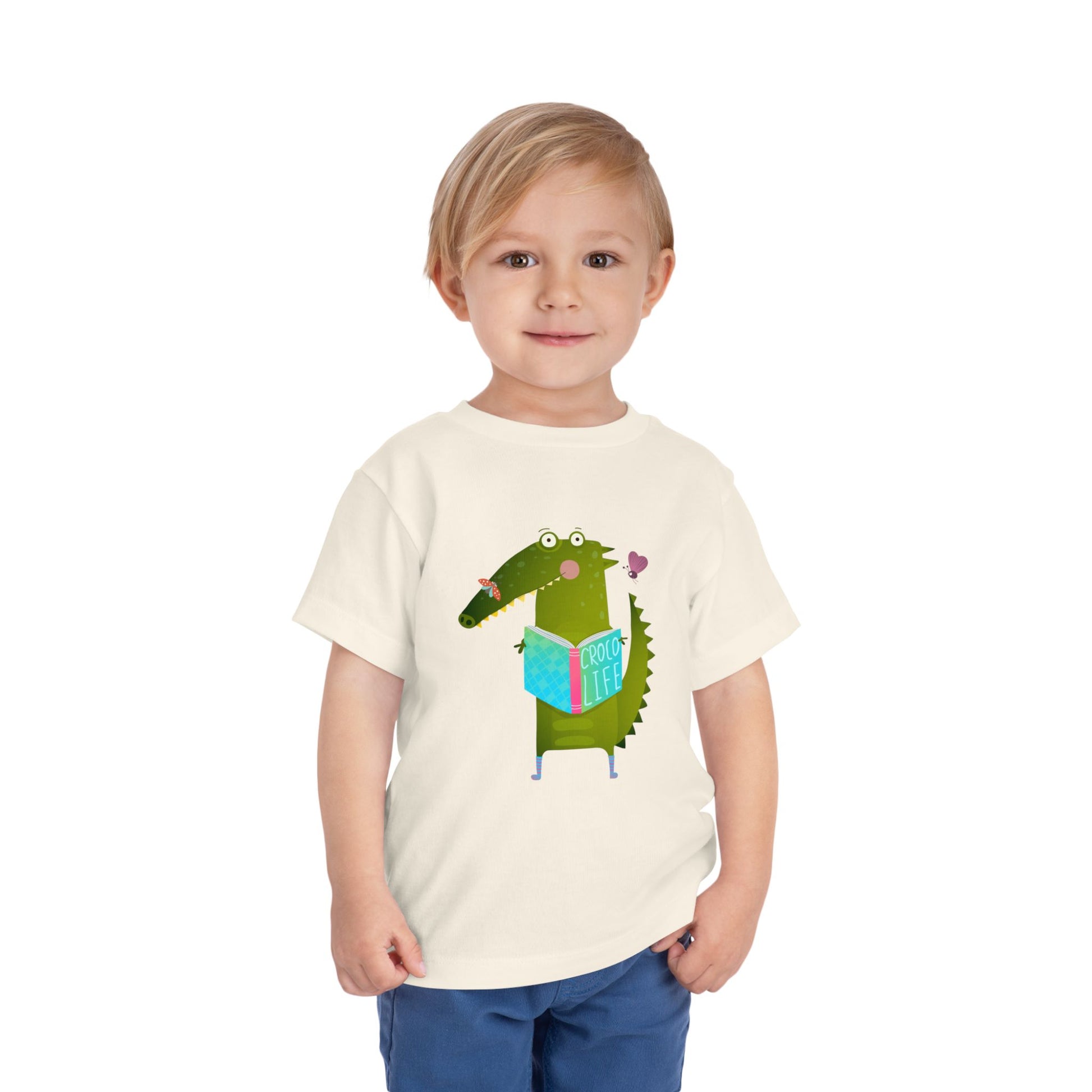 Toddler wearing a beige t-shirt with a cartoon crocodile reading a Croc Life book amongst sweet insects 