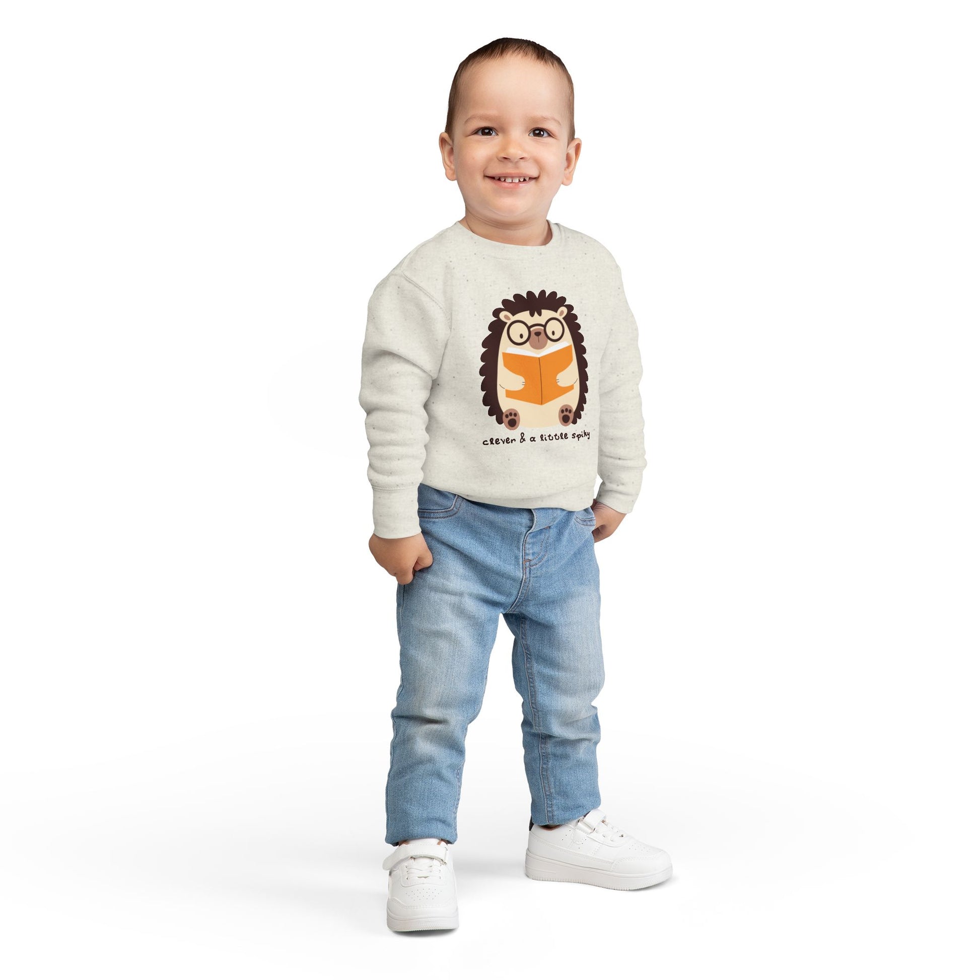 Boy wearing a beige toddler sweatshirt with a hedgehog graphic reading a book and text 'clever & a little spiky'