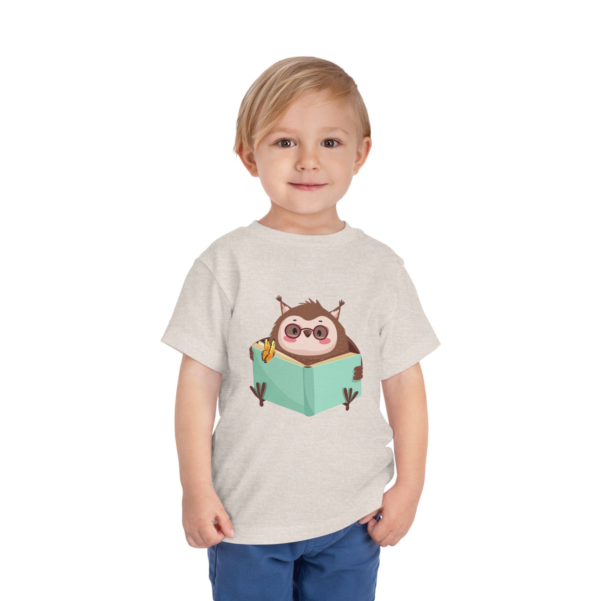 Child wearing a beige toddler t-shirt with a cartoon own reading a book with a butterfly perched on top
