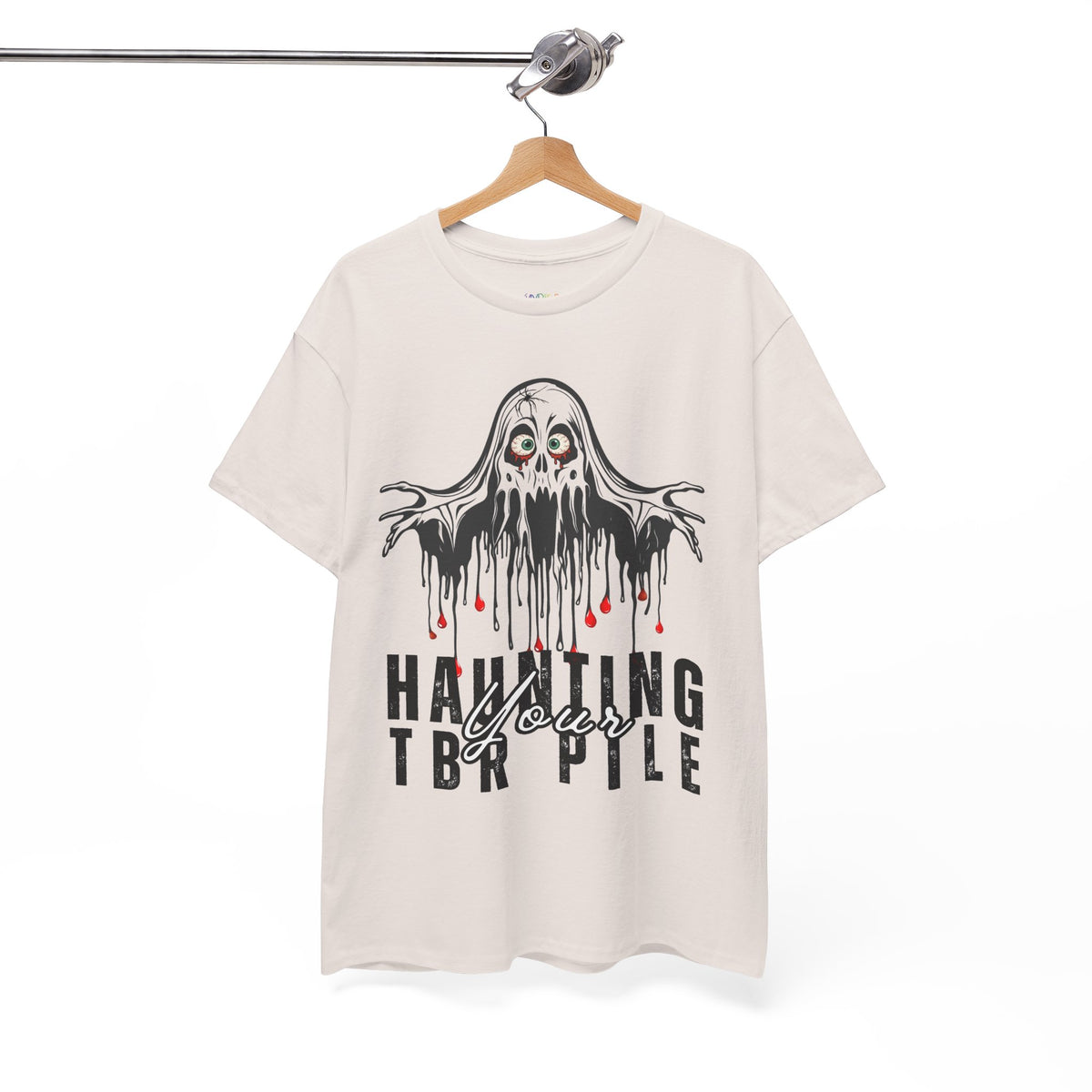 A tan short-sleeve t-shirt hanging on a wooden hanger from a metal rack. The shirt features a graphic of a dripping black ghost with red blood-like drips and the text 'HAUNTING YOUR TBR PILE' printed below it.