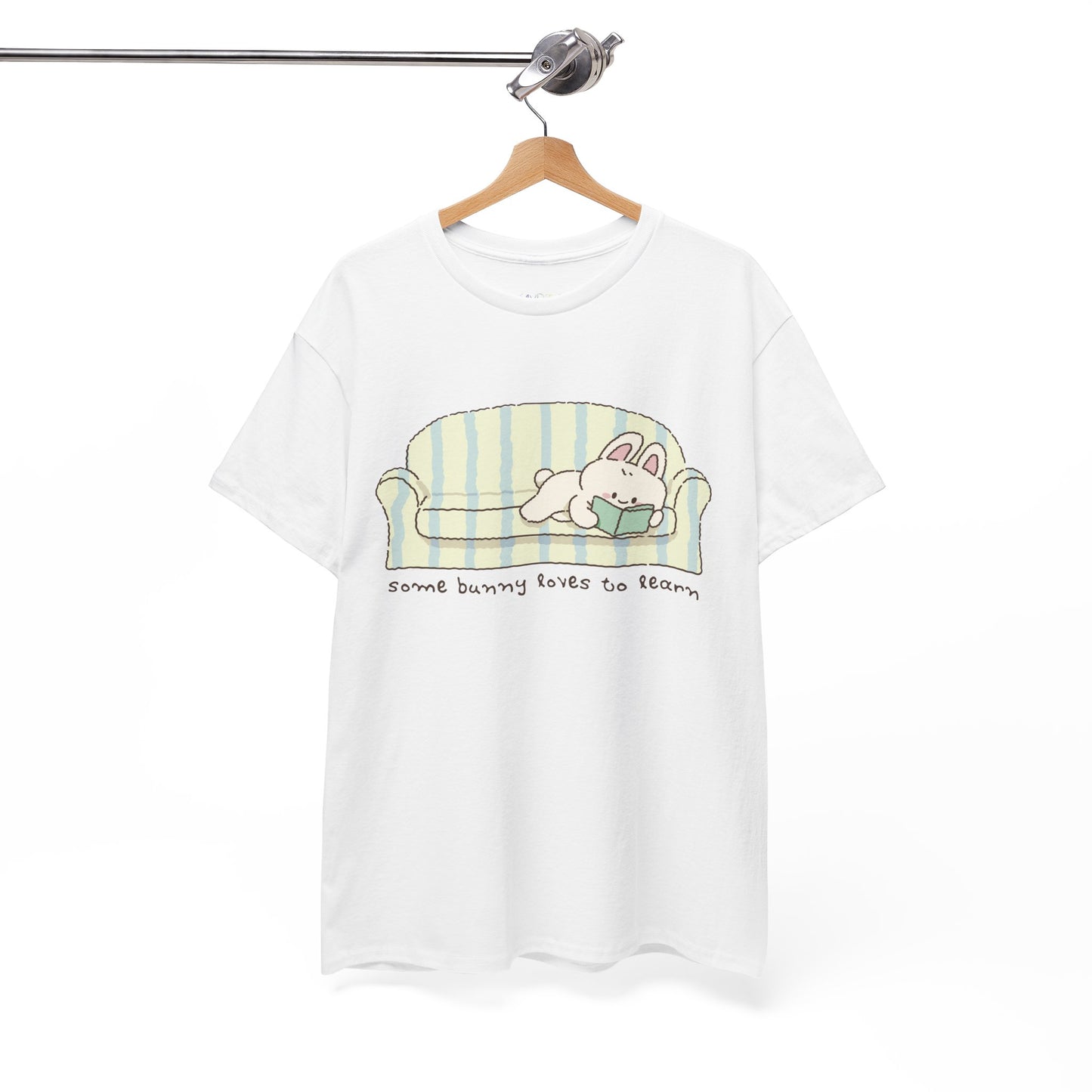 White t-shirt with a cartoon bunny reading a book as it's lays on a couch, text 'some bunny loves to learn' 