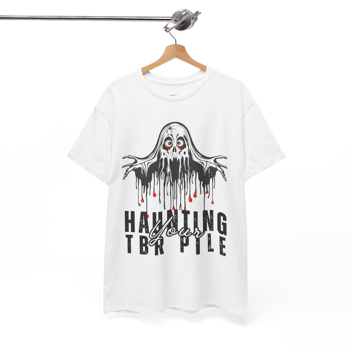 A white short-sleeve t-shirt hanging on a wooden hanger from a metal rail, featuring a black dripping ghost graphic with red eyes and the text 'HAUNTING YOUR TBR PILE' in black and white with small red accents.