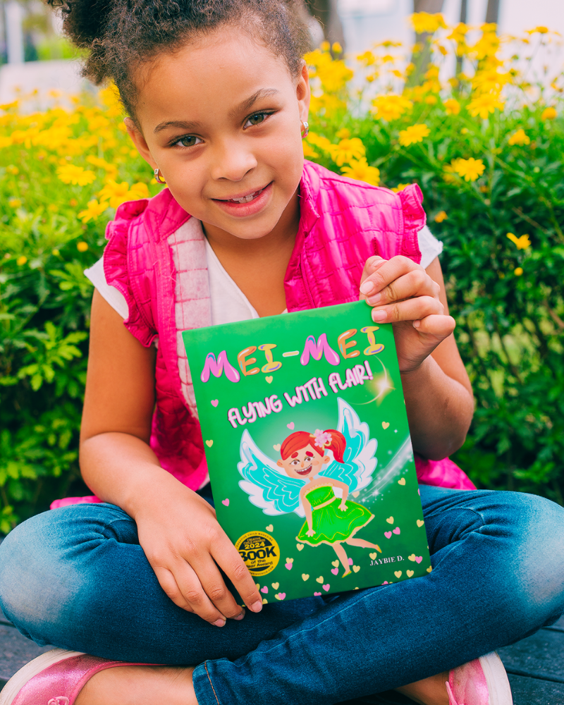 Child holding a book titled 'MEI-MEI Flying with Flair' in a garden setting