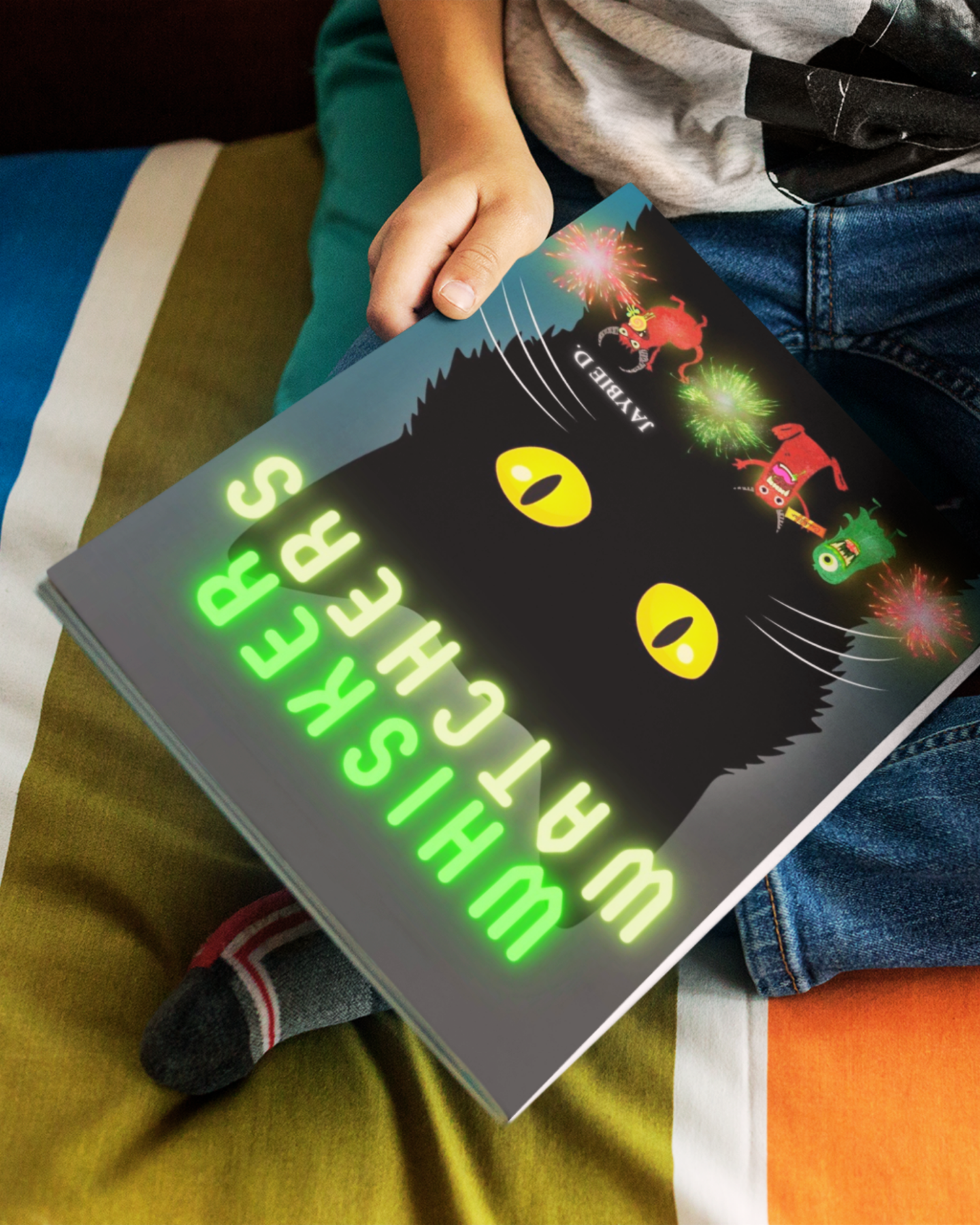 Child holding a the children's picture book 'Whisker Watchers" by Jaybie D. upside down in his lap as he sits on a colorful comforter.