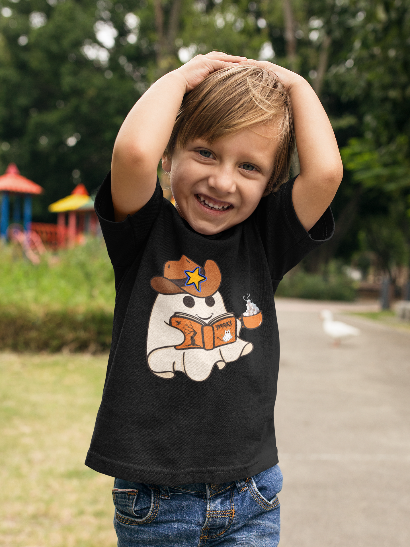 Toddler boy wearing a black t-shirt featuring a cowboy hat wearing ghost who is reading a book and holding a cup of hot chocolate with a pile of marshmallows.