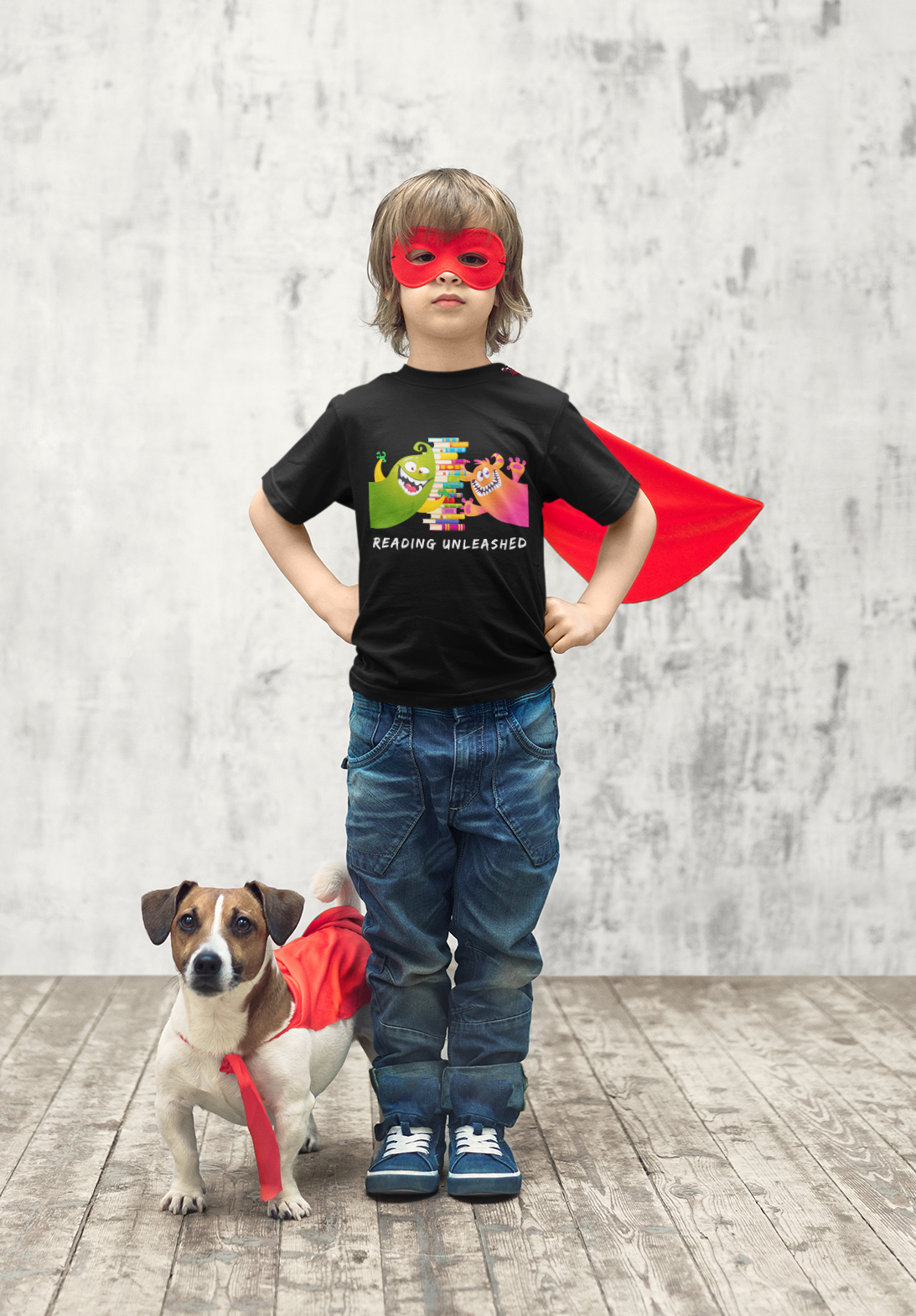Little boy wearing a mask, cape and monster t-shirt next to his dog wearing a cape.
