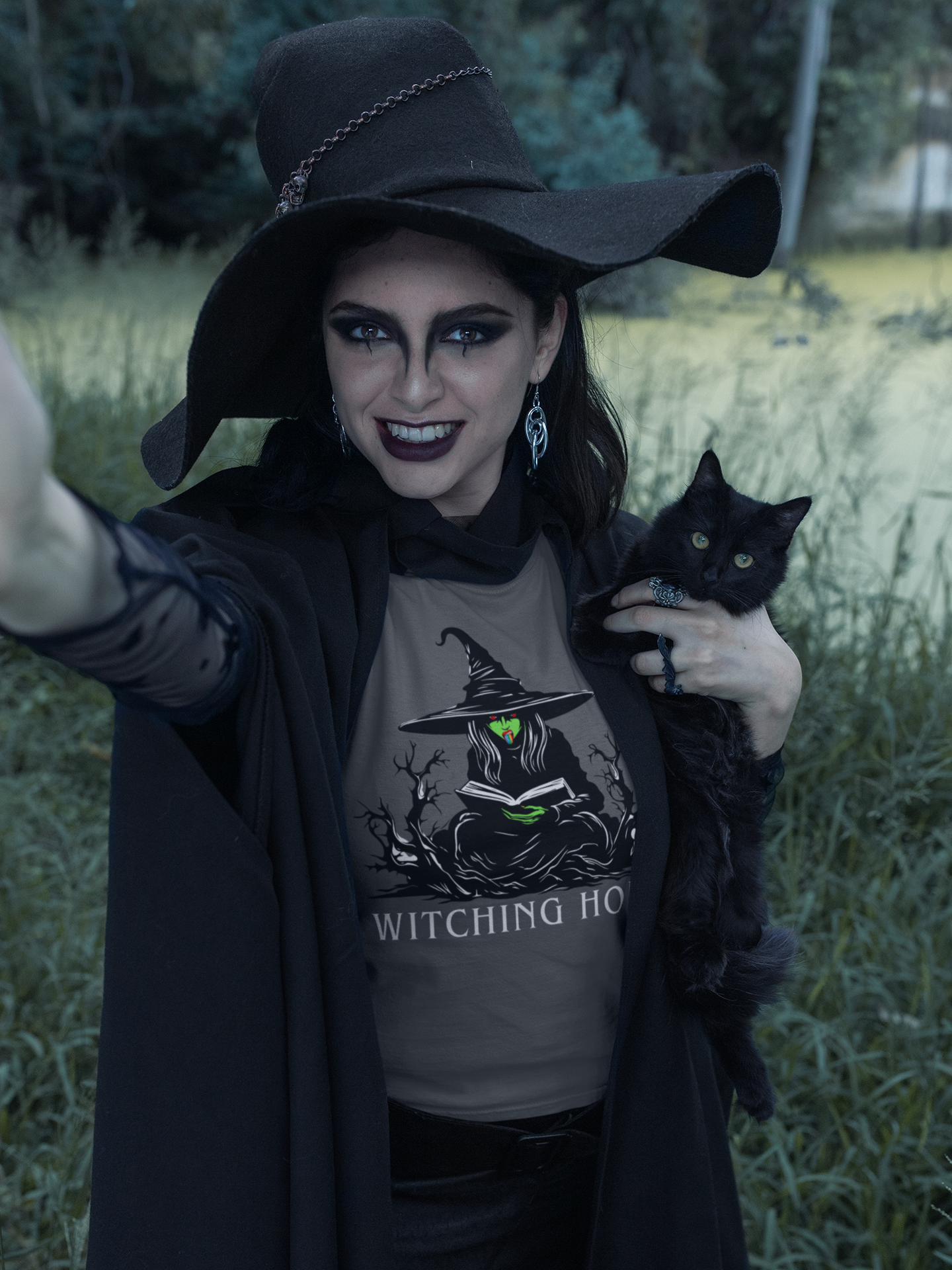 A witch in a dusk setting, holding a black cat and wearing a "Witching Hour" t-shirt featuring a green witch sticking out a rainbow tongue.